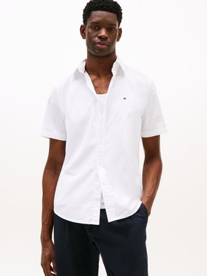 Jovem vestido com uma camisa de botão branca de mangas curtas sobre uma camisola branca, com calças escuras e uma mão no bolso.