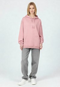 Nő áll, rózsaszín, túlméretezett kapucnis pulóvert, laza szürke farmert és fehér platform tornacipőt viselve, sima világos háttér előtt.