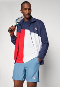 Tumesinine ja valge treeningjakk punaste detailidega, koos helesiniste lühikesed pükstega. Omab Fila logo, sportlikku disaini ja siledat tekstuuri.