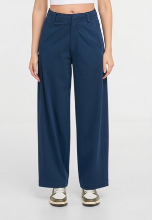 Frau trägt marineblaue, weit geschnittene Hose mit hoher Taille und weiße Sneaker und steht vor einem schlichten weißen Hintergrund.