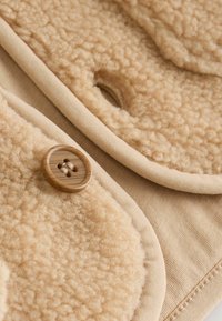 Primo piano di un tessuto in pile beige con texture, caratterizzato da un grande bottone marrone e cuciture rinforzate dell'asola.