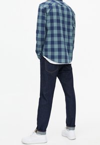 Camisa de cuadros azul y verde, vaqueros de mezclilla oscuros y zapatillas blancas. La camisa tiene un dobladillo redondeado y botones en los puños.