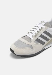 Ljust grå Adidas-sneaker med vita snören, mesh- och mockadetaljer, grå ränder och svart sula på en vit bakgrund.