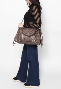 Grande borsa grigia in suede con parte superiore arrotondata, molteplici tasche con cerniera e dettagli decorativi in metallo. La borsa presenta frange e una tracolla lunga.