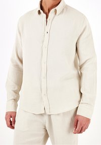 Camicia button-up beige chiaro in tessuto strutturato, con colletto e maniche lunghe, bottoni visibili e una vestibilità comoda.