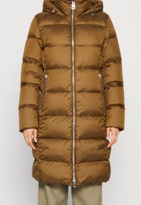 Lange bruine gewatteerde winterjas met een rits aan de voorkant, twee met rits gesloten zijzakken en een capuchon, gedragen over beige broek.