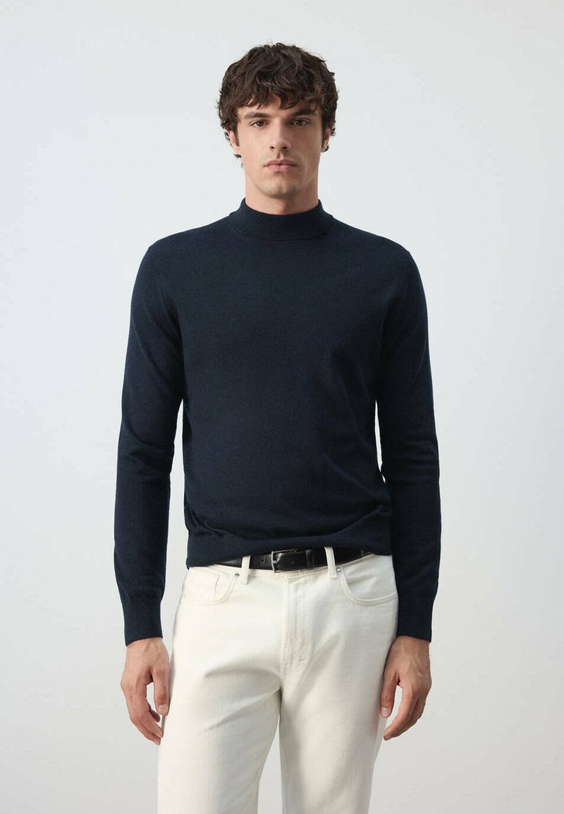 Jeune homme debout, portant un pull à col roulé bleu marine foncé, un pantalon blanc et une ceinture noire sur un fond clair uni.