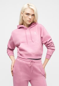 Νεαρή γυναίκα με ξανθά μαλλιά, φορώντας ροζ φούτερ Converse με κουκούλα και αντίστοιχο παντελόνι φόρμας, στέκεται με τα χέρια στη μέση μπροστά σε λευκό φόντο.