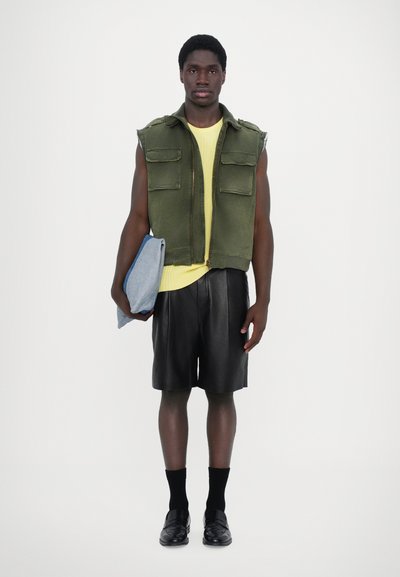 Veste sans manches verte sur un pull en tricot jaune, short en cuir noir, chaussettes noires et mocassins, tenant un vêtement en denim bleu.