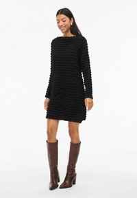 Kvinna som bär en texturerad svart långärmad knälång klänning och höga bruna klackstövlar, leende med händerna avslappnat vid sidorna.