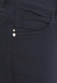 Donkerblauwe denim spijkerbroek heeft een voorzak met twee zilveren hardwareaccenten. Glad textuur met subtiele stikdetails.