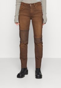 Pantalones de mezclilla marrón con un diseño ajustado, parches en las rodillas, una cinturilla de altura media y texturas contrastantes. Terminados con detalles cosidos.