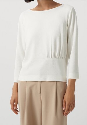 Femme portant un haut blanc à manches longues avec encolure bateau et un pantalon plissé beige, les mains détendues le long du corps sur un fond uni.