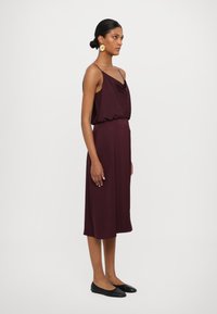 Rochie burgundă cu bretele subțiri, bodice drapat, talie elastică și fustă până la genunchi; asortată cu balerini negri și cercei aurii.