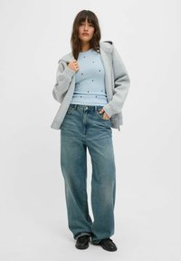 Šedá zipová mikina přes světle modré vzorované tričko a volné světle modré džíny. Černé boty dotvářejí outfit. Jednoduchý design.