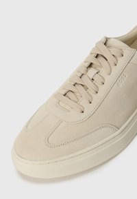 Beige suede sneaker with flat laces, rounded toe, and a minimalist design. Features a stitched outline and subtle branding on the side.