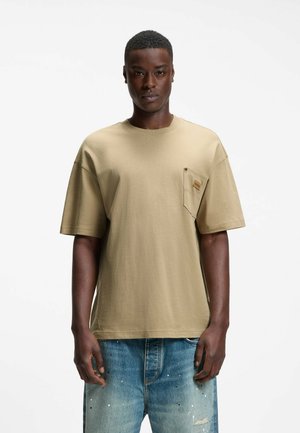 Homme portant un T-shirt beige à manches courtes avec une poche poitrine et un jean bleu usé sur un fond blanc uni.