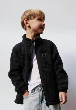 Gutt iført svart fleecejakke med glidelås, hvit skjorte og lyseblå jeans, ser til siden med en hånd i lomma.