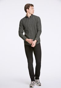Camisa de botones verde oscuro con una tela texturada, mangas largas y un corte entallado, combinada con pantalones negros de corte slim y zapatillas.