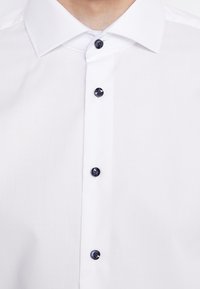 Vit skjorta med strukturerad krage och fem svarta knappar framtill, slät tyg med en subtil lyster.