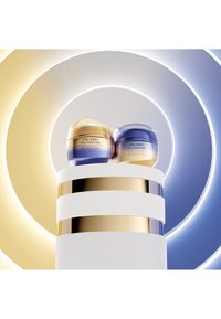 Två burkar av Shiseido Vital Perfection kräm, en guld och en blå, placerade på ett vitt och guldrandigt podium, mot cirkulära ljusbakgrunder.