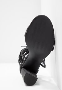 Chaussure noire à talons hauts avec tissu texturé, une large sangle et une semelle en caoutchouc lisse. Le talon se rétrécit vers le haut avec une face avant plate.