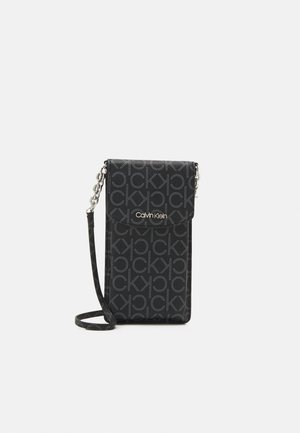 Petit sac bandoulière noir Calvin Klein avec motif répétitif du logo CK et chaîne argentée sur fond blanc.