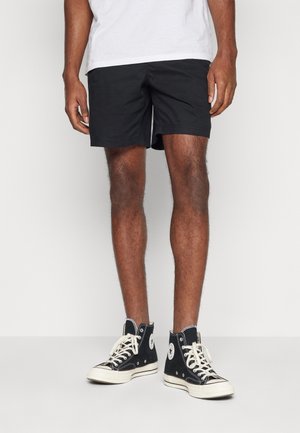 Hombre con pantalones cortos negros, zapatillas altas negras con cordones blancos y camisa blanca, de pie frente a un fondo liso.