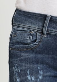 Vaqueros de mezclilla azul con un acabado desgastado, con acentos desgastados, un pequeño bolsillo frontal y un logo "RAW" en la cinturilla.