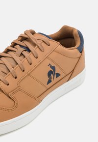 Sneaker in pelle marrone chiaro con lacci piatti, dettagli blu navy, un logo sul lato e suola in gomma bianca. Presenta una texture liscia e un design classico.