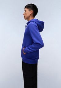 Felpa blu in cotone con cappuccio regolabile, tasca frontale e polsini a costine. Abbinata a pantaloni cargo neri, mostrata di profilo.