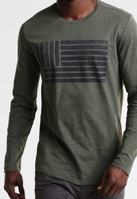 Langsleeve shirt in olijfgroen met een zwart grafisch patroon van horizontale strepen die op een vlag lijken. Heeft een ronde halslijn en een gestructureerde stof.