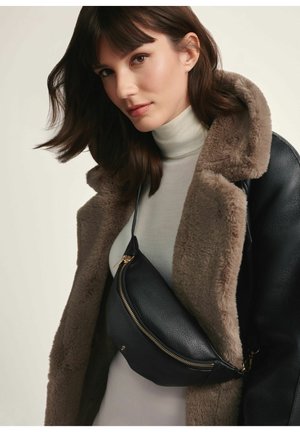 Czarna skórzana nerka o zaokrąglonym kształcie, złoty zamek błyskawiczny i regulowany pasek, noszona na kremowym golfie i brązowej kurtce z futrzanym podszyciem.