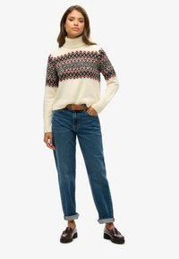 Crème turtleneck sweater met een rood en zwart geometrisch patroon, gecombineerd met blauwe rechte jeans en donkere loafers.