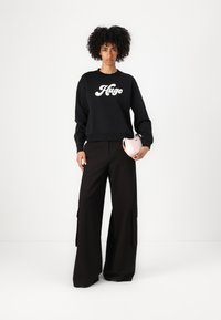 Felpa nera con logo bianco "Hugo", abbinata a pantaloni cargo neri a gamba larga con tasche laterali e una piccola borsa rosa.