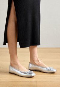 Ballet flats argentati con punta rotonda, dettaglio a fiocco e finitura metallica liscia. Indossati con un vestito nero a coste su un pavimento in legno.