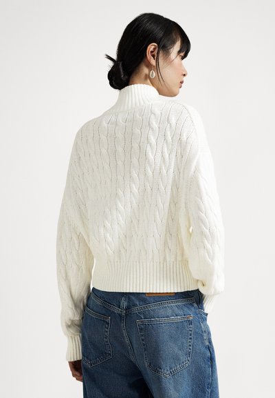 Γυναίκα με σκούρο μαλλί σε κότσο που φοράει λευκό πουλόβερ με ζιβάγκο και μοτίβο πλεκτού καλωδίου, καθώς και μπλε τζιν, απεικονίζεται από πίσω με απλό φόντο.
