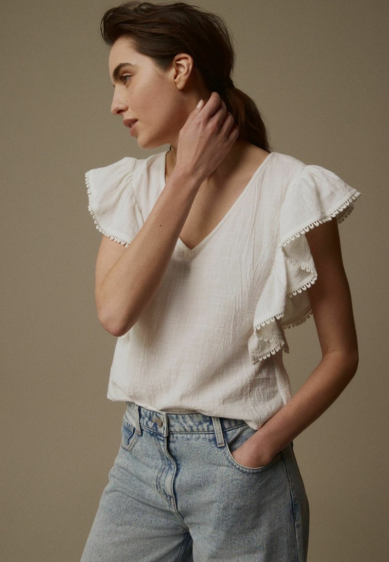 Blouse blanche à manches courtes avec des manches volantes et une finition en dentelle, associée à un jean taille haute bleu clair. Tissu doux au tombé décontracté.