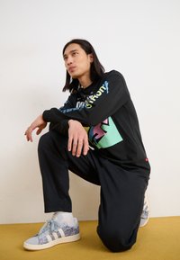 Black graphic sweatshirt with colorful prints, black pants, and light blue sneakers with a pattern. Model is crouching on a yellow surface.