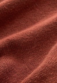 Lähikuva pehmeästä, ruosteenvärisestä neuloksesta, jossa näkyvät tekstuuriset silmukat ja lempeät laskokset.