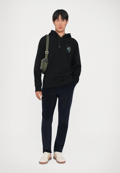 Čierna mikina s kapucňou a farebnou výšivkou králika, v kombinácii s námorníckymi nohavicami a béžovými teniskami. Olivová crossbody taška.