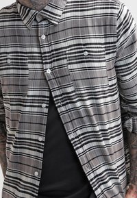 Camicia di flanella a quadri grigia e nera con colletto a punta, due tasche sul petto e una consistenza morbida. Indossata sopra una t-shirt nera.