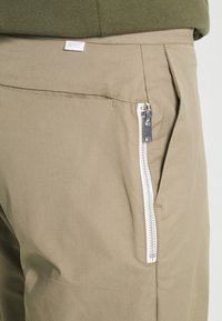 Béžové kalhoty Nike s elegantní boční kapsou na zip, hladkou strukturou a moderním designem, který zdůrazňuje střih na míru a minimální akcenty.