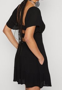 Kvinna med långa flätor som bär en kort svart klänning med öppna ryggband, korta ärmar och en hand i fickan.