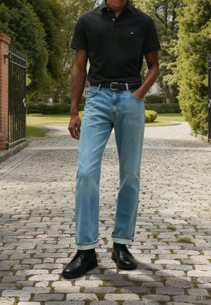 Mann trägt schwarzes Poloshirt, hellblaue Jeans mit hochgekrempelten Bündchen, schwarzen Gürtel und schwarze Lederschuhe, steht auf einem Kopfsteinpflasterweg im Freien.