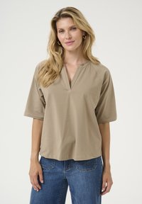 Top beige de manga corta con escote en forma de V y mangas raglán. Está hecho de una tela suave y lisa. Combinado con unos jeans de mezclilla azul.