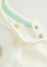 Trumpomis rankovėmis kremo spalvos palaidinė su turkio spalvos apykakle, puošta geltonos saulės ir vaivorykštės siuvinėjimu bei sidabriniais spaustukais ant peties.