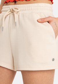 Cream-colored shorts made of soft fabric with an elastic waistband, drawstring, side pockets, and a small branded metal accent.