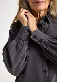 Grå denimskjorta med knapprad, plisserade ärmar, krage och struktur. Närbild som fokuserar på manschetterna och tyget.