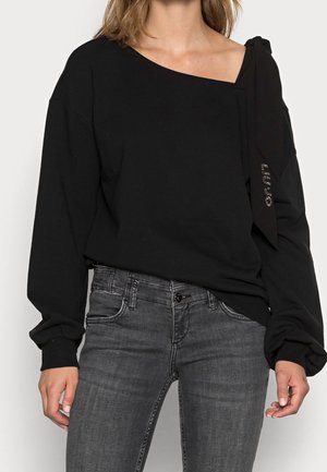 Woman wearing black off-shoulder sweater with long puffed sleeves and dark grey fitted jeans, standing against a plain background.